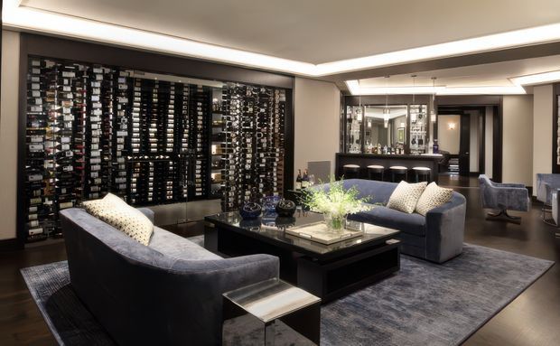 A sophisticated, spacious living room with dark grey upholstered sofas, patterned cushions, a large low black coffee table, and a textured area rug. A dramatic full-height wine display wall occupies the left side, while a mirrored bar or entertaining zone is visible at the rear. Recessed perimeter ceiling lighting, dark timber-toned flooring, and a neutral charcoal, black, and warm grey palette create a refined, moody atmosphere.