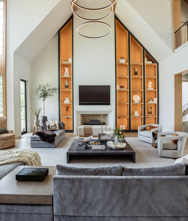 A spacious, refined living room with a dramatic double-height vaulted ceiling and a central fireplace wall. Tall built-in timber display shelving frames the fireplace and television, while multiple grey upholstered sofas and armchairs surround a dark rectangular coffee table. Large windows and glazed doors provide abundant natural light, and sculptural ring pendant lights hang overhead. A stair and upper-level balustrade are visible at the right.