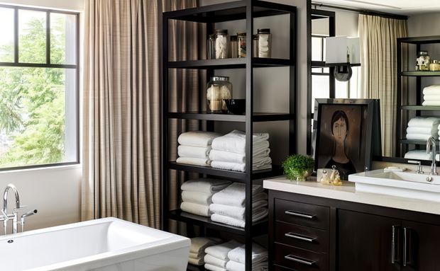 A refined bathroom with a freestanding white bathtub beside a large window, dark wood vanity cabinetry, a white countertop with a vessel sink, brushed metal tapware, and an open black-framed shelving unit displaying neatly folded towels and bathroom accessories. Full-height striped curtains, mirrors, and greenery add softness and visual depth.