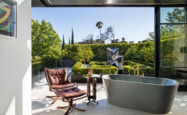 A bright contemporary bathroom opening directly onto a landscaped garden terrace through large black-framed glazing. The room features a freestanding dark grey bathtub, a leather lounge chair with ottoman, a small side table, pale flooring, and a sculptural outdoor artwork visible beyond the glazing.