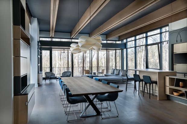 Open-plan contemporary living and dining space with a long timber dining table, dark upholstered dining chairs, a lounge seating area, and a compact kitchen or bar zone. Full-height black-framed glazing overlooks a wooded setting. Exposed timber ceiling beams contrast with dark ceiling panels, while built-in timber and dark cabinetry line the walls. A sculptural white pendant light hangs above the dining table.