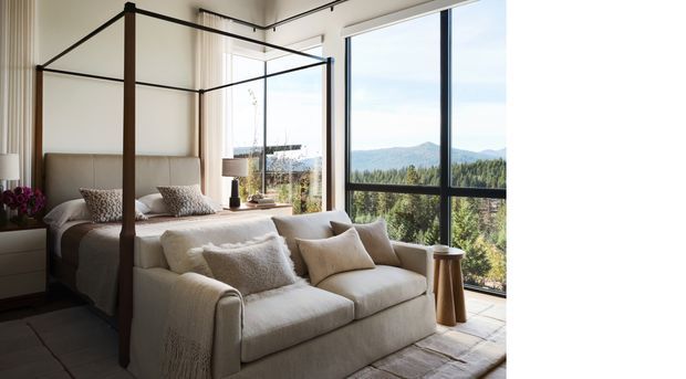 A bright, refined bedroom with a four-poster bed positioned beside full-height black-framed glazing and a cream upholstered sofa at the foot of the bed. Neutral textiles, dark flooring, simple bedside furnishings, and a mountain-and-forest outlook create a calm, contemporary retreat.