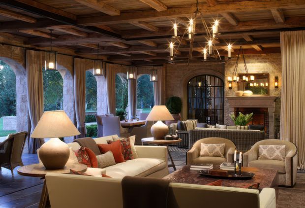 A richly layered lounge or sitting room with exposed dark timber ceiling beams, rustic stone walls, tall arched windows, and a large arched black-framed doorway. The room is furnished with cream sofas and armchairs, patterned and rust-coloured cushions, timber side tables, table lamps, and a substantial dark wood coffee table. Warm pendant and chandelier lighting creates an inviting, lodge-like atmosphere.