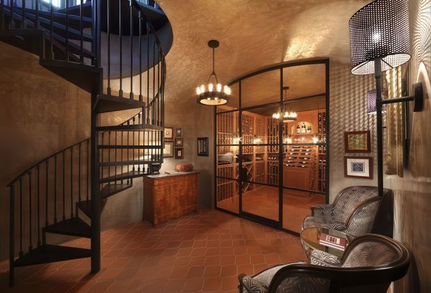 Warm, richly detailed interior featuring a sculptural black metal spiral stairway, terracotta tiled floor, textured earthen-toned walls and ceiling, and a glazed black-framed enclosure revealing a wine storage and tasting area. Decorative framed artwork, patterned lounge chairs, timber cabinetry, and pendant and wall lighting create an intimate, luxurious atmosphere.