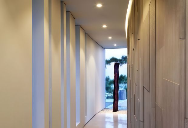 A long, narrow contemporary hallway with a glossy light floor, smooth pale walls, recessed ceiling downlights, and tall vertical wall panels or built-in doors along the right side. Slender structural or decorative vertical strips line the left wall, creating a rhythmic corridor. At the far end, full-height glazing or an opening reveals an outdoor area and a tall sculptural feature. The restrained palette and concealed detailing create a refined, gallery-like atmosphere.