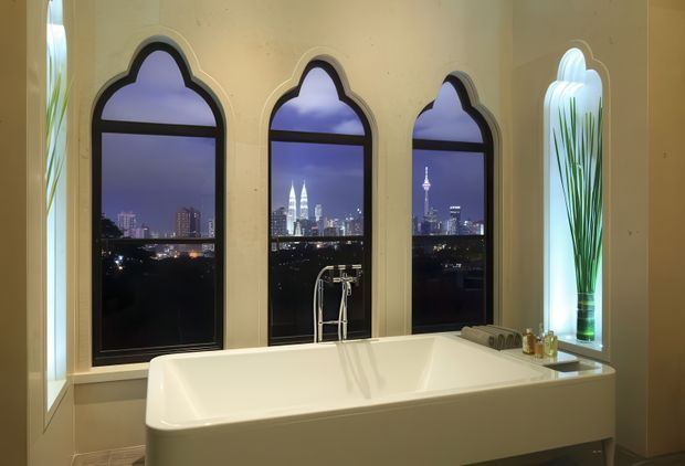 Elegant bathroom featuring a freestanding white bathtub positioned before three tall, pointed ogee-arched windows overlooking a city skyline at dusk. The warm cream walls, dark window frames, sculptural arched side glazing, and subtle integrated lighting create an ornate, luxurious atmosphere.