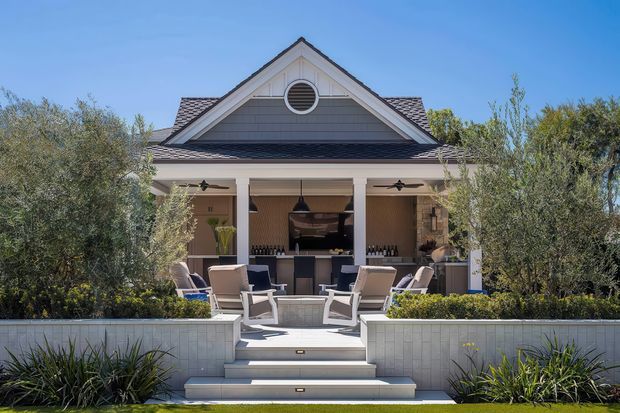A symmetrical exterior view of a refined gable-roofed outdoor entertaining pavilion. The structure has a light rendered facade, dark shingle-style roof, circular attic window, covered veranda with white columns, and an outdoor kitchen or bar at the rear. White steps lead to a paved terrace furnished with lounge chairs, surrounded by landscaped planting and low rendered retaining walls.