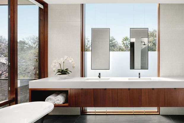 A refined contemporary bathroom vanity with a long floating dark timber cabinet, white integrated benchtop and twin basins. Two rectangular mirrors sit against vertically ribbed pale wall panels, with warm timber-framed full-height glazing on the left and frosted lower glazing behind the vanity. A freestanding white bath is partially visible in the foreground, and the room is finished with dark linear floor tiles and minimal black fixtures.