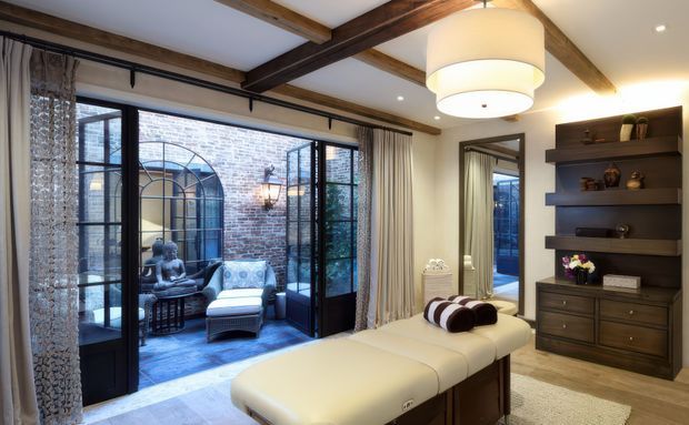 Elegant contemporary sitting room with exposed dark timber ceiling beams, large black-framed glazed doors opening to a brick courtyard, a cream upholstered chaise lounge, patterned drapery, and dark built-in shelving and cabinetry. The room is warmly lit and combines refined contemporary furnishings with traditional architectural detailing.
