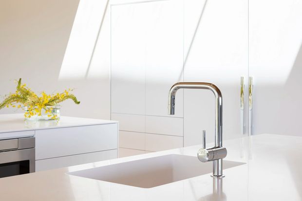 Bright minimalist kitchen with white handleless cabinetry, a large pale island benchtop and integrated sink, plus a prominent chrome gooseneck mixer tap. A built-in oven is partly visible at left, with a simple vase of yellow flowers adding a small accent. Strong daylight enters through large angled roof glazing or skylights.