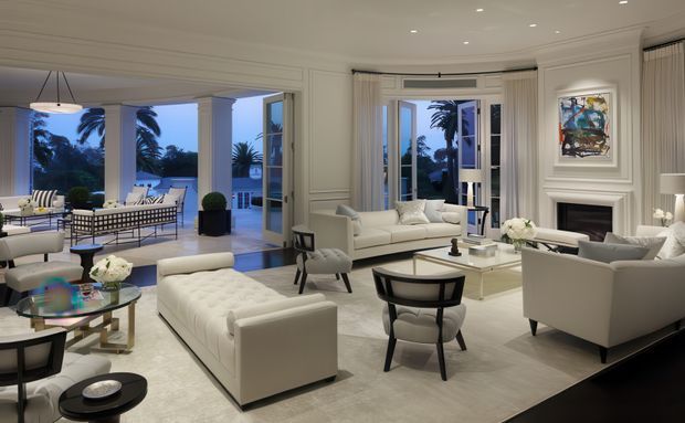 A refined, spacious living room with white paneled walls, ornate fireplace mantel, recessed ceiling lighting, and large French doors opening to a covered outdoor terrace. The room is furnished with cream sofas, a tufted ottoman, glass coffee tables, and dark accent chairs, creating a bright formal interior with views of tropical landscaping and water beyond.