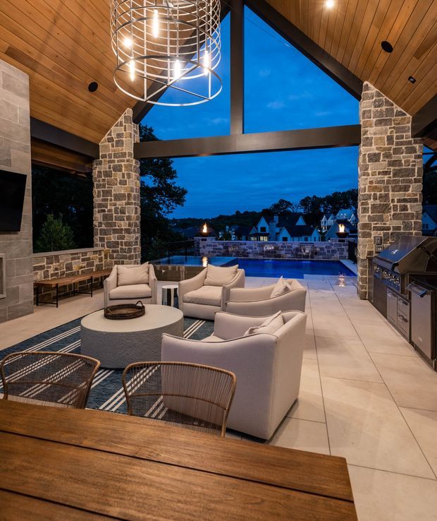 A sophisticated covered outdoor entertaining area opening onto a swimming pool and waterfront or lakeside view at dusk. The space features a timber-lined vaulted ceiling, exposed dark structural beams, large triangular openings, substantial irregular-stone masonry columns and walls, pale tiled flooring, upholstered lounge seating around a round fire-table, wicker dining chairs and a large timber dining table. A built-in stainless-steel outdoor kitchen and grill occupies the right side, while a sculptural circular metal pendant hangs overhead. The atmosphere is warm, luxurious and inviting.