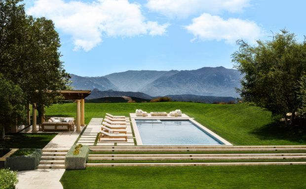 A landscaped outdoor pool terrace with a rectangular swimming pool, broad stone steps, timber-and-stone retaining edges, sun loungers, and a covered outdoor seating pavilion. Manicured lawns and mature trees frame the scene, with dramatic mountain ranges visible beyond the property under a bright blue sky.