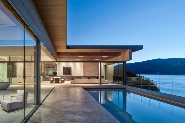A contemporary waterfront residence viewed from an outdoor terrace, with expansive floor-to-ceiling glazing opening onto a refined living area. The home features strong horizontal roof overhangs, warm timber soffits, dark framed glass, concrete paving, and an infinity-edge swimming pool overlooking calm blue water and a wooded hillside.