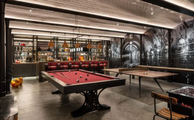 A dramatic industrial-style games and entertainment room featuring a red-felt billiards table in the foreground, a ping-pong table to the right, and a fully stocked backlit home bar with pendant lights and red bar stools. Dark graphic wall murals, black metal framing, exposed ceiling services, and polished concrete flooring create a moody urban atmosphere.