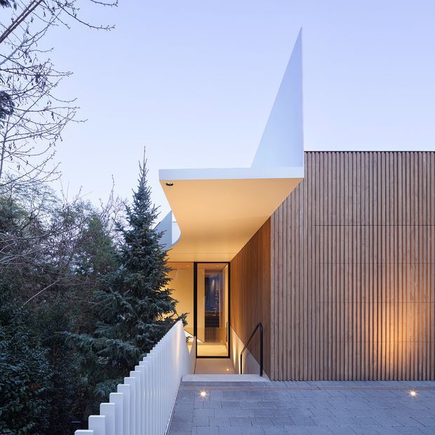Exterior architectural view of a contemporary residence entry, featuring a sharply cantilevered white roof canopy, warm vertical timber cladding, glazed entry door, illuminated stair access, and a paved terrace. Mature evergreen planting partially screens the building.