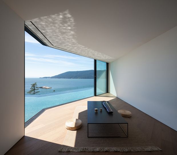 Minimalist contemporary living room with a low dark coffee table and floor rug, opening through expansive corner glazing onto a pale timber deck and an infinity pool overlooking calm coastal water and a forested headland. White walls and ceiling, dark timber flooring, black-framed glazing, and strong patterned sunlight create a serene, refined interior with a dramatic view.