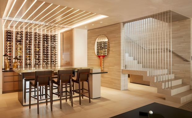 A refined contemporary residential wine room and entertaining area with extensive illuminated wine display racks, a compact bar island with dark timber stools, warm timber wall panelling, and a sculptural open staircase screened by slender vertical metal balusters. Integrated linear ceiling lighting and neutral stone-look flooring create a warm, luxurious atmosphere.