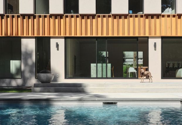 Contemporary two-storey residential facade viewed across a swimming pool. The building combines dark-framed glazing and pale rendered walls with a prominent upper-level timber screen of vertical slats. A broad concrete terrace and steps lead to the pool, with large sliding glass openings revealing a sparsely furnished interior. The composition is clean-lined and strongly horizontal, with warm timber contrasting against a monochrome facade.