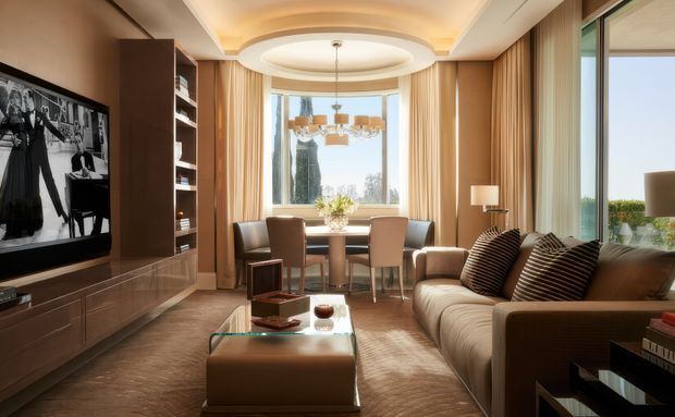 A refined open-plan living and dining room with a warm neutral palette. The room features a large upholstered sofa, glass coffee table, built-in television and cabinetry, a dining table positioned beside expansive glazing, layered curtains, and a sculptural recessed ceiling with concealed lighting. The space feels polished, bright, and luxurious, with views to greenery beyond the windows.