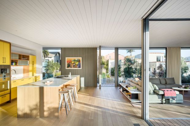 Bright open-plan interior combining a yellow kitchen with a central island and stools, a dining area, and a living room visible through large glazed sliding doors. The space features extensive timber flooring, a vertical timber feature wall, white-lined ceiling, abundant daylight, and strong indoor-outdoor visual connection.