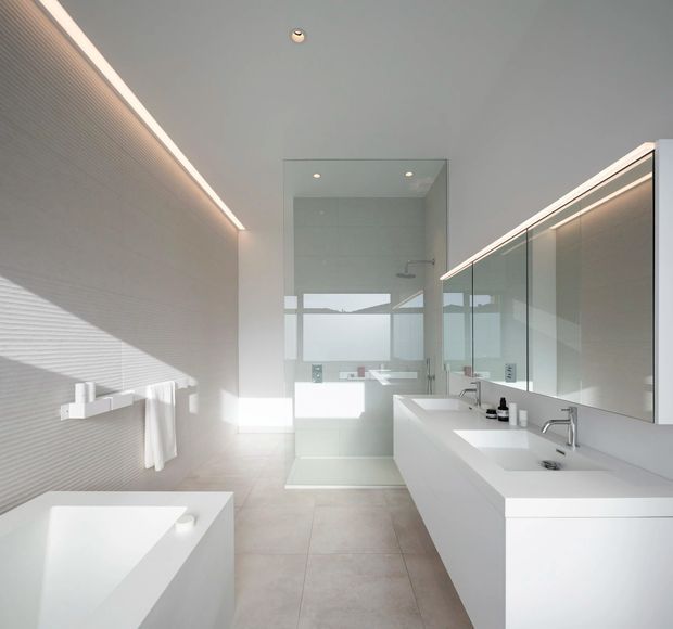Bright, spacious contemporary bathroom with a freestanding white bathtub in the foreground, a large double vanity with integrated basins and wall-to-wall mirrored cabinetry, and a frameless glass shower enclosure at the rear. Pale large-format floor tiles, fine linear wall tiling, concealed storage, and integrated linear lighting create a clean, minimalist appearance. A window or glazed opening provides strong natural light and exterior views.