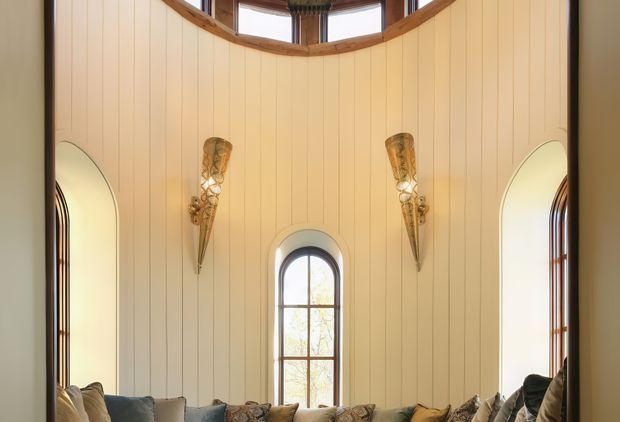 A tall, curved sitting-room interior with cream vertical wall panelling, a high arched window, and matching narrow arched side openings. Warm timber trim follows the upper curved glazing, while ornate wall sconces and the tops of a sofa with cushions create a refined, dramatic atmosphere.