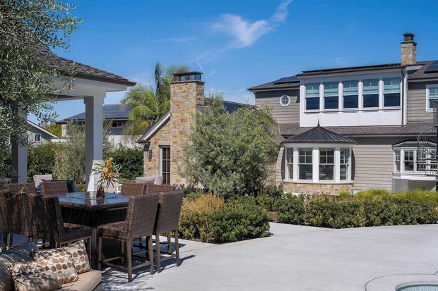 Sunny landscaped residential courtyard viewed from an outdoor dining terrace. The scene features a substantial multi-wing home with grey shingle-style cladding, white-trimmed windows, stone chimney, dark hipped roof sections, a covered outdoor dining area, paved entertaining space, planting, and a partial glimpse of a pool at the lower right.