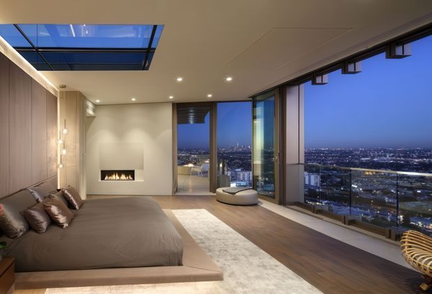 A luxurious high-rise bedroom at dusk, featuring a low platform bed, warm wood wall panelling, a recessed linear fireplace, expansive floor-to-ceiling glazing, and a glass skylight. The room overlooks a brightly lit urban cityscape, with a neutral palette and integrated recessed lighting.