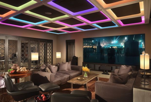 A richly furnished home media room with multiple sofas and lounge chairs arranged around a large wall-mounted screen. The room features a dramatic geometric recessed ceiling with multicoloured LED lighting, dark acoustic ceiling panels, patterned decorative screens or cabinetry along the left wall, table lamps, a central stone or wood coffee table, and warm brown and taupe furnishings. The atmosphere is cinematic, luxurious, and moody.