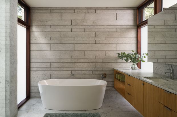 A refined bathroom with a freestanding white bathtub positioned against a full-height horizontal grey block wall. Dark timber-framed corner glazing brings in natural light, while a timber vanity with integrated drawers, open shelving, and a pale stone benchtop runs along the right side beneath a large mirror. The room has a restrained, modern material palette with concrete-like grey surfaces, warm timber joinery, and minimal styling.