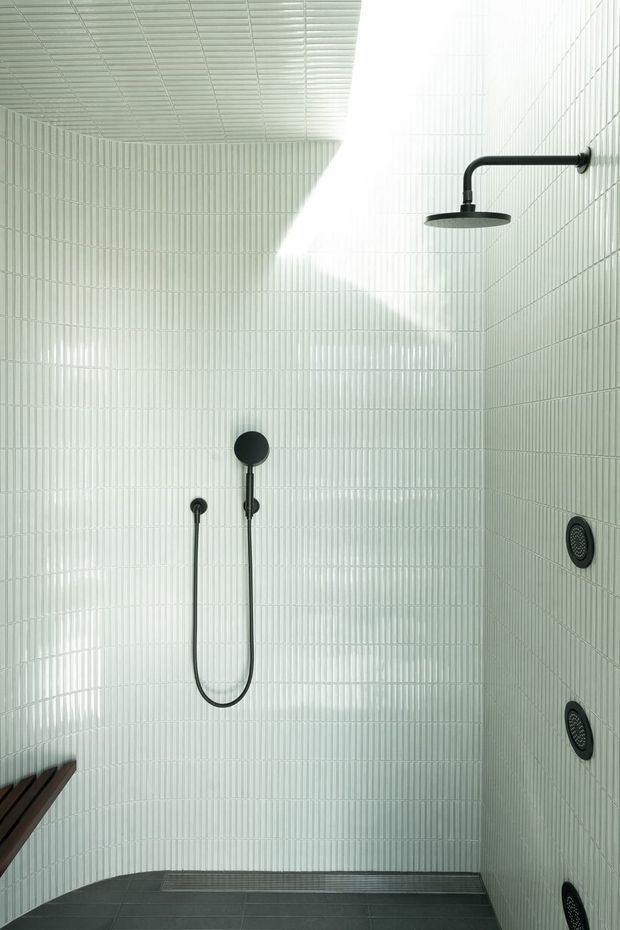Bright, minimalist walk-in shower lined floor-to-ceiling with narrow vertically ribbed white tiles. Matte black shower fittings include a ceiling-mounted rain shower, handheld shower, controls, and multiple wall jets. The floor is finished in dark grey tiles, creating strong contrast with the pale walls.