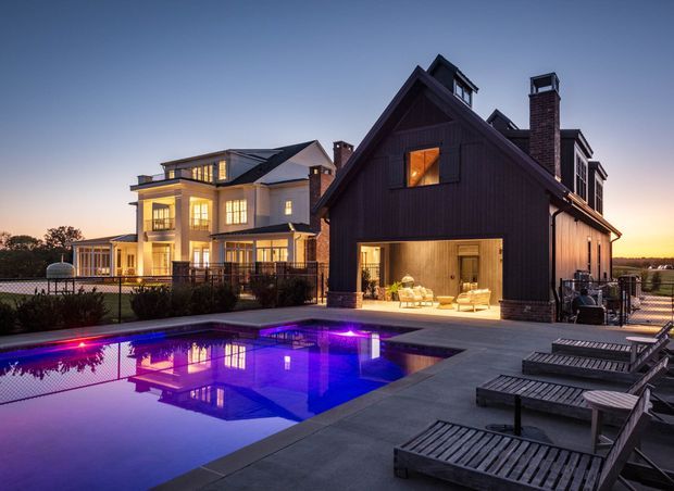 Exterior dusk view of a large residential property with a dark vertical-clad gabled pavilion, adjacent light-coloured multi-level house, expansive glazed openings, and a glowing covered outdoor living area. A swimming pool with vivid purple-blue lighting occupies the foreground, surrounded by concrete paving and timber sun loungers. The scene has a refined resort-like atmosphere and appears to be a multi-storey suburban or rural estate.