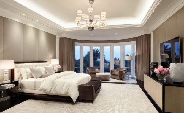 Luxurious, spacious bedroom with a large upholstered bed, padded feature wall, dark timber bedside tables and bench, neutral carpet, full-height curved glazing, layered drapery, ornate chandelier, and built-in media cabinetry. The room opens visually toward an exterior balcony or terrace through the glazing.
