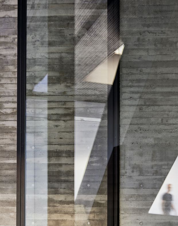 Close-up architectural view of a dark grey board-formed concrete facade with tall black-framed glazing. Strong geometric reflections and angular sunlight create sharp light-and-shadow patterns across the concrete and glass; a faint human silhouette is visible in reflection.