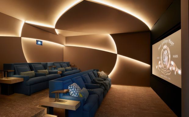 A dark, luxurious home cinema with tiered rows of deep blue reclining lounge seating, small wooden side tables, and a large projection screen. Sculptural curved wall and ceiling panels are outlined with warm concealed LED lighting, creating an immersive theatrical atmosphere. The floor is carpeted in a warm brown tone.
