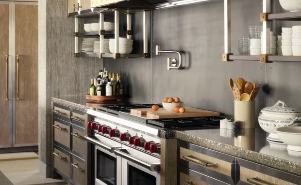 A refined, traditional-style kitchen focused on a large professional range with red controls, stainless-steel appliances, dark metal open shelving, and extensive grey stone benchtops. The space features dark grey textured wall finishes, metallic cabinetry with brass-toned hardware, timber accents, and open shelving displaying crockery and glassware.