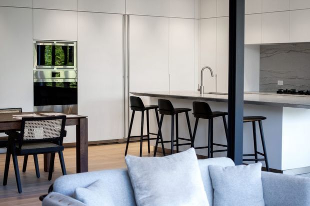 Open-plan contemporary kitchen and dining area viewed from a living room. The kitchen features floor-to-ceiling white flat-panel cabinetry, a long stone island with integrated seating, a stainless-steel tap, built-in appliances, and a grey marble-look splashback. A dark timber dining table and black woven dining chairs sit beside the kitchen, while a pale upholstered sofa occupies the foreground. The space is bright, refined, and minimalist with strong black-and-white contrast and warm timber flooring.