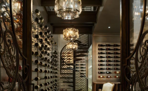 A richly detailed, dark-toned wine cellar or wine room viewed through ornate decorative metal-and-glass doors. Extensive wall-mounted wine bottle displays line both sides, with a sculptural spiral staircase at the rear, dark timber ceiling beams, reflective glass, and multiple ornate chandelier-style pendant lights. The space has a luxurious, dramatic, highly crafted appearance.