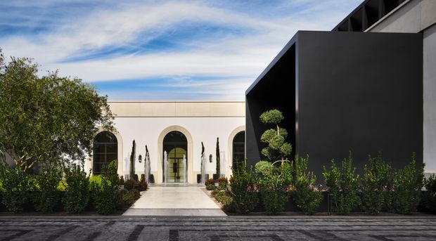 Exterior architectural view of a refined modern residence or estate, combining a dark angular flat-roofed volume with a pale symmetrical entrance pavilion featuring tall arched windows and doors. A formal landscaped garden, clipped topiary, cypress-like planting, paved walkway, and dark foreground paving reinforce the structured composition.