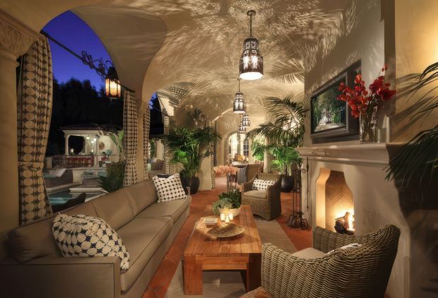 An atmospheric, richly decorated living room opening onto an outdoor area through large arched openings. The room features a long upholstered sofa, woven rattan armchairs, a substantial timber coffee table, patterned curtains and cushions, indoor palms, ornate hanging lanterns casting dappled shadows across the curved ceiling, and a traditional fireplace with a lit fire. Warm terracotta flooring and cream walls create an inviting, resort-like ambience.