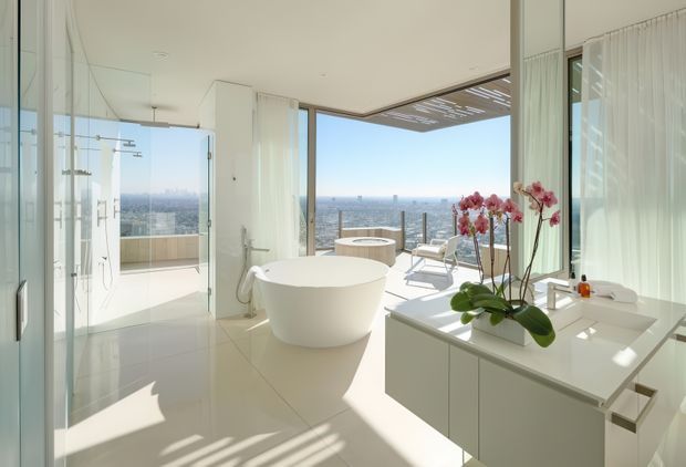 A bright, luxurious contemporary bathroom with a freestanding white bathtub, frameless glass shower enclosure, and minimalist white vanity cabinetry with an integrated white benchtop. Full-height glazing and an open terrace provide expansive coastal or city views, while sheer curtains and an orchid add softness. The space has a clean, airy, high-end atmosphere with abundant natural light.