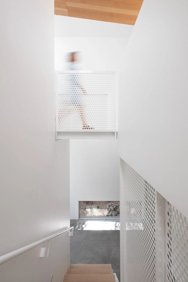 A minimalist interior stairway viewed from above, with pale timber stair treads descending toward a polished grey floor and a small window at the landing. White walls and a perforated white metal balustrade create a light, airy enclosure. A timber-lined ceiling section adds warmth, while a perforated artwork or screen is visible on the upper wall.