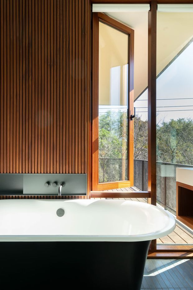 A refined contemporary bathroom centered on a freestanding white bathtub with a dark exterior. The room features a warm vertical timber slat feature wall, integrated dark grey bath fittings, timber-framed glazing, and a partially open door leading to a balcony or terrace with views of trees. Natural light and the elevated outlook create an airy, warm atmosphere.