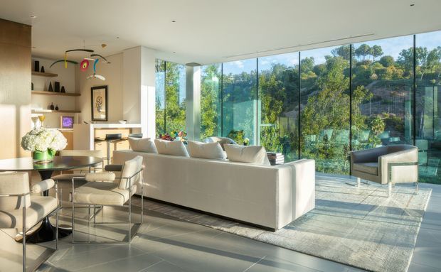 Bright, open-plan contemporary living and dining area with a large white sectional sofa, glass coffee table, dining table and upholstered dining chairs. Floor-to-ceiling glazing overlooks a leafy landscape, while warm timber joinery and open shelving line the left side. The room has a minimalist, light-toned interior with strong natural light and a refined indoor-outdoor connection.