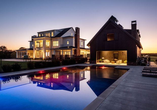 Twilight exterior view of a large residential property with two adjacent buildings beside a reflective swimming pool. The main house is a light-coloured, multi-storey traditional-style residence with extensive windows, a pitched roof, chimneys, and an illuminated covered terrace. A darker timber-clad gabled structure sits alongside it, featuring a large open garage or entertaining bay with visible dining furniture. The pool and paved deck dominate the foreground, with warm interior lighting reflected in the water.