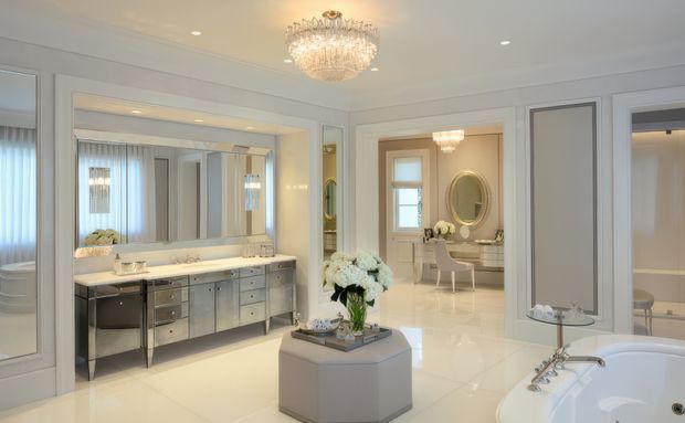 Luxurious, bright bathroom interior with a large double vanity, mirrored wall, polished light flooring, freestanding bathtub in the foreground, and an adjoining elegant dressing or bedroom area visible through the opening. Crystal chandeliers, pale neutral finishes, metallic cabinetry, and symmetrical detailing create a refined classic atmosphere.