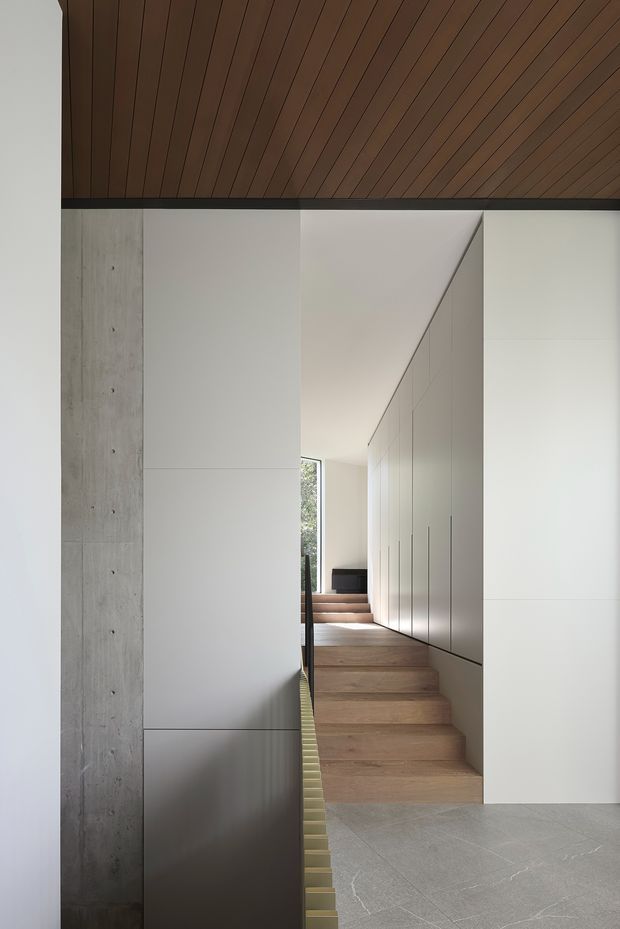 Minimalist interior corridor and stairway with warm timber slatted ceiling, pale grey-white full-height built-in cabinetry, exposed raw concrete wall, timber stairs, and large-format light grey floor tiles. The narrow stair passage leads toward a softly lit window and dark console, creating a clean, refined architectural composition.