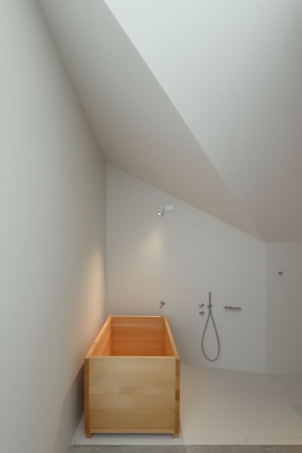 Minimalist bathroom with a freestanding light timber soaking tub positioned against a plain white wall. A wall-mounted shower with handheld hose, simple tapware, recessed lighting, and a partially sloped ceiling create a quiet, spa-like interior. The room has a restrained palette of white, pale wood, and light grey flooring.