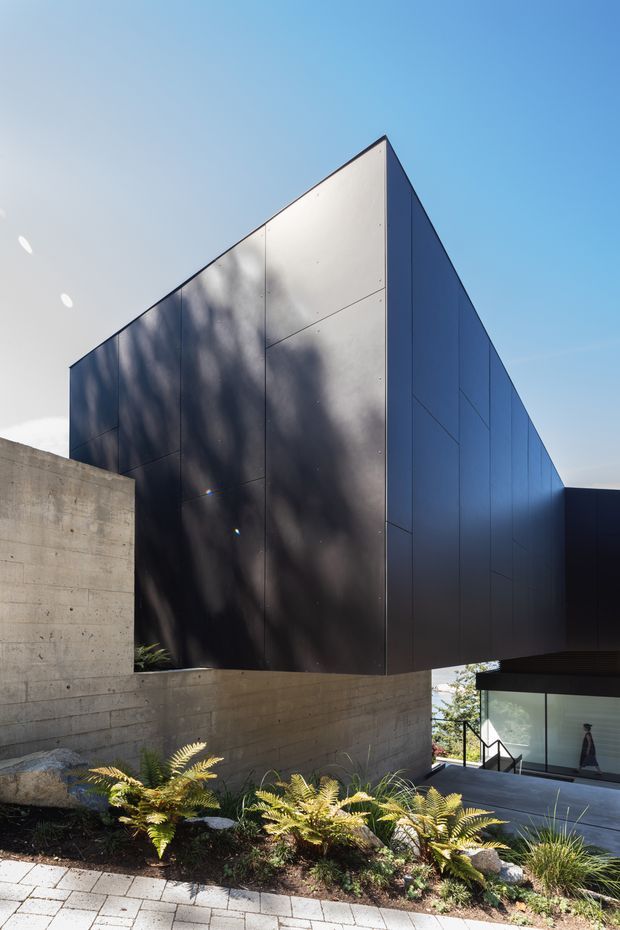 Exterior architectural photograph of a sharply angular, wedge-shaped building volume clad in dark charcoal metal panels. The structure rises above exposed concrete retaining walls on a sloped landscaped site, with a glazed lower-level entrance or living area visible at right. The composition emphasizes the faceted geometric form, crisp panel joints, and strong contrast against a clear blue sky.
